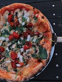 A freshly baked pizza topped with tomatoes, mushrooms, spinach, and onions. The crust appears to be crispy and brown, and the pizza is served on a metallic platter. The background is a dark wooden surface.