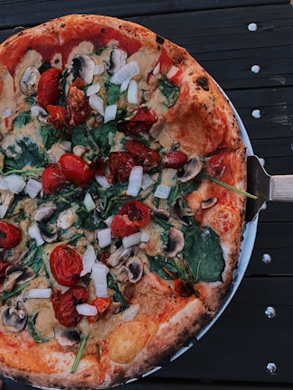 A freshly baked pizza topped with tomatoes, mushrooms, spinach, and onions. The crust appears to be crispy and brown, and the pizza is served on a metallic platter. The background is a dark wooden surface.