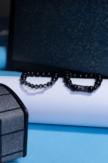 Two black beaded bracelets are placed on a white cylindrical object against a backdrop of a dark textured surface and a light blue platform. The bracelet on the left spells out a message with lettered beads.