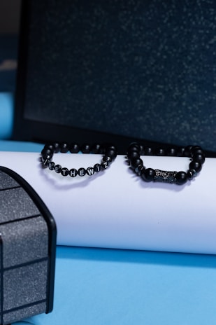Two black beaded bracelets are placed on a white cylindrical object against a backdrop of a dark textured surface and a light blue platform. The bracelet on the left spells out a message with lettered beads.