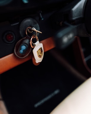 A set of car keys hanging in the ignition of a car, with a leather keychain displaying a Porsche emblem. The dashboard surface is visible, featuring a wooden trim and buttons, creating a vintage or classic car aesthetic.
