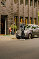 A person wearing sunglasses and a hat stands on a street next to a parked Fiat 500. The individual is dressed casually in a denim jacket and sneakers. The backdrop features a brick building with large windows and a sign reading 'Cotélac'. A potted plant is noticeable near the entrance.