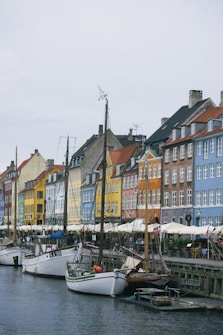 Brightly colored buildings line a waterfront street, showcasing shades of yellow, red, blue, and orange. In front of the buildings, several sailboats with tall masts are docked along a canal, adding a maritime charm. People can be seen enjoying outdoor seating under white umbrellas, giving the scene a lively, casual atmosphere.