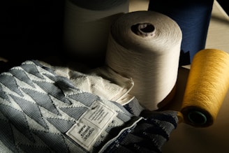A collection of large spools of thread in various colors, including cream, navy, and yellow, are arranged beside a fabric with a geometric pattern. The fabric has a label that reads 'www.towel.studio' and some text in German. The lighting is dramatic, casting shadows across the scene, highlighting the textures of the threads and fabric.