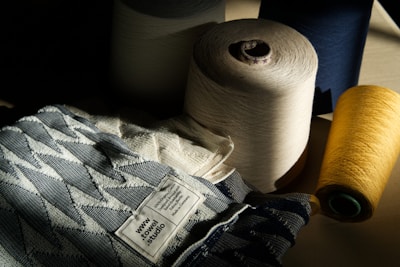 A collection of large spools of thread in various colors, including cream, navy, and yellow, are arranged beside a fabric with a geometric pattern. The fabric has a label that reads 'www.towel.studio' and some text in German. The lighting is dramatic, casting shadows across the scene, highlighting the textures of the threads and fabric.