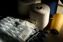 A collection of large spools of thread in various colors, including cream, navy, and yellow, are arranged beside a fabric with a geometric pattern. The fabric has a label that reads 'www.towel.studio' and some text in German. The lighting is dramatic, casting shadows across the scene, highlighting the textures of the threads and fabric.