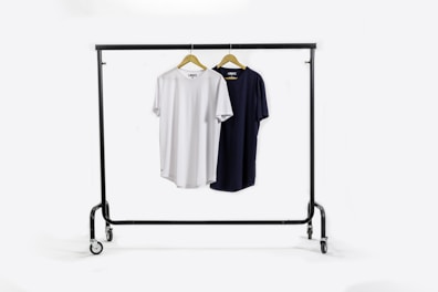 A clothing rack holds two t-shirts on wooden hangers. The t-shirt on the left is white, and the one on the right is black. The rack has wheels for mobility and stands against a plain white background.