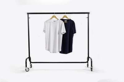 A clothing rack holds two t-shirts on wooden hangers. The t-shirt on the left is white, and the one on the right is black. The rack has wheels for mobility and stands against a plain white background.