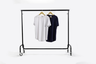 A clothing rack holds two t-shirts on wooden hangers. The t-shirt on the left is white, and the one on the right is black. The rack has wheels for mobility and stands against a plain white background.