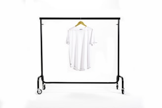 A white T-shirt hangs centrally on a wooden hanger suspended from a black metal clothing rack with wheels. The background is a clean white, creating a minimalist and uncluttered appearance.