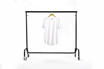 A white T-shirt hangs centrally on a wooden hanger suspended from a black metal clothing rack with wheels. The background is a clean white, creating a minimalist and uncluttered appearance.