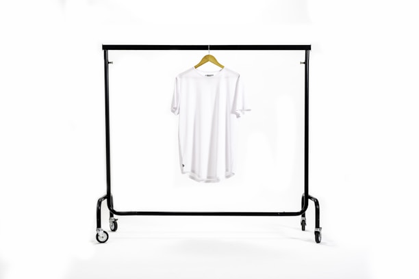 A white T-shirt hangs centrally on a wooden hanger suspended from a black metal clothing rack with wheels. The background is a clean white, creating a minimalist and uncluttered appearance.