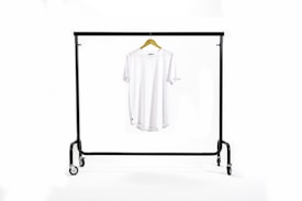 A white T-shirt hangs centrally on a wooden hanger suspended from a black metal clothing rack with wheels. The background is a clean white, creating a minimalist and uncluttered appearance.