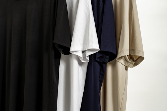 Several T-shirts hang vertically, featuring different colors including black, white, navy blue, and beige. The T-shirts are displayed side by side, showcasing their short sleeves and simple fabric texture.