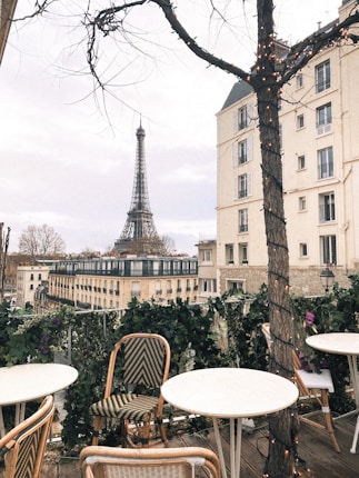 A charming outdoor caf&eacute; with empty tables and chairs, framed by a tree with lights wrapped around its trunk. In the background, the Eiffel Tower stands tall amidst Parisian buildings under a cloudy sky.