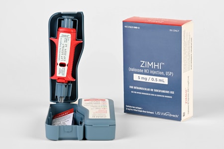 A naloxone injection device and its packaging are displayed. The device is red with a gray base and housed in a gray plastic case. The packaging is a blue and white box with branding and dosage information prominently visible. Additional instructional materials are included inside the case.