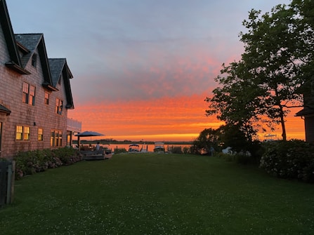 A tranquil lakeside scene during sunset with vibrant orange and pink hues in the sky. A large house with multiple windows and a peaked roof stands on the left, while an outdoor seating area with a table and umbrella is situated on the patio. To the right, a lush green lawn and trees frame the view, leading to a calm, reflective body of water.