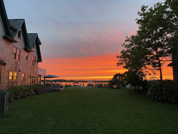 A tranquil lakeside scene during sunset with vibrant orange and pink hues in the sky. A large house with multiple windows and a peaked roof stands on the left, while an outdoor seating area with a table and umbrella is situated on the patio. To the right, a lush green lawn and trees frame the view, leading to a calm, reflective body of water.