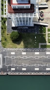 An aerial view of a tall building with a red and white rooftop standing beside a green lawn. The building casts a shadow on the lawn, which is adjacent to a paved area with square patterns and street lamps. A curved structure and additional paved areas are visible to the side. The edge of a body of water is seen at the bottom of the image.