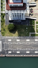 An aerial view of a tall building with a red and white rooftop standing beside a green lawn. The building casts a shadow on the lawn, which is adjacent to a paved area with square patterns and street lamps. A curved structure and additional paved areas are visible to the side. The edge of a body of water is seen at the bottom of the image.