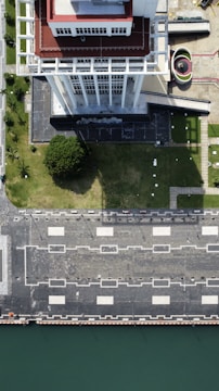 An aerial view of a tall building with a red and white rooftop standing beside a green lawn. The building casts a shadow on the lawn, which is adjacent to a paved area with square patterns and street lamps. A curved structure and additional paved areas are visible to the side. The edge of a body of water is seen at the bottom of the image.