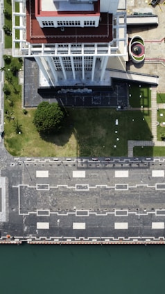 An aerial view of a tall building with a red and white rooftop standing beside a green lawn. The building casts a shadow on the lawn, which is adjacent to a paved area with square patterns and street lamps. A curved structure and additional paved areas are visible to the side. The edge of a body of water is seen at the bottom of the image.