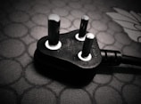 A close-up view of a black electrical plug with three prongs resting on a textured surface. The plug features metallic prongs and engraved markings indicating voltage and current specifications.