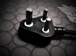 A close-up view of a black electrical plug with three prongs resting on a textured surface. The plug features metallic prongs and engraved markings indicating voltage and current specifications.