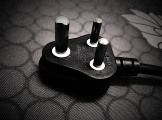 A close-up view of a black electrical plug with three prongs resting on a textured surface. The plug features metallic prongs and engraved markings indicating voltage and current specifications.