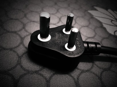 A close-up view of a black electrical plug with three prongs resting on a textured surface. The plug features metallic prongs and engraved markings indicating voltage and current specifications.