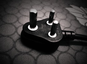 A close-up view of a black electrical plug with three prongs resting on a textured surface. The plug features metallic prongs and engraved markings indicating voltage and current specifications.