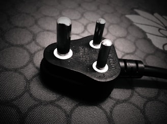 A close-up view of a black electrical plug with three prongs resting on a textured surface. The plug features metallic prongs and engraved markings indicating voltage and current specifications.