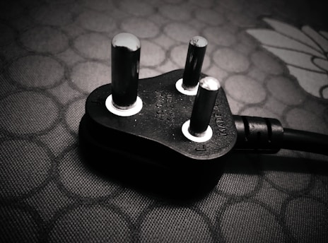 A close-up view of a black electrical plug with three prongs resting on a textured surface. The plug features metallic prongs and engraved markings indicating voltage and current specifications.