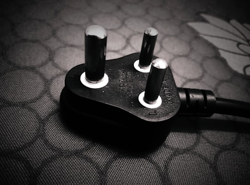 A close-up view of a black electrical plug with three prongs resting on a textured surface. The plug features metallic prongs and engraved markings indicating voltage and current specifications.
