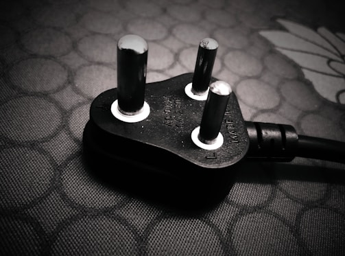 A close-up view of a black electrical plug with three prongs resting on a textured surface. The plug features metallic prongs and engraved markings indicating voltage and current specifications.