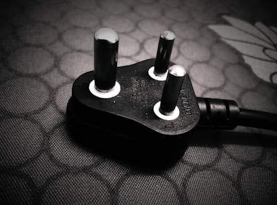 A close-up view of a black electrical plug with three prongs resting on a textured surface. The plug features metallic prongs and engraved markings indicating voltage and current specifications.