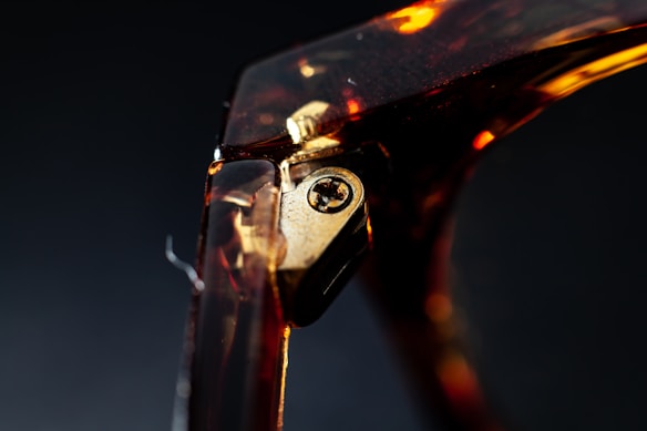 A close-up view of a tortoiseshell eyeglass hinge, featuring a metallic screw. The image captures the intricate details and textures of the frame in low light conditions, highlighting the reflective quality of the materials.