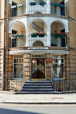 A multi-story building with arched balconies containing flower pots with red flowers, black railings, and a grand entrance with a staircase leading up to double glass doors. The building is made of light-colored brick and features a sign reading 'Mall Chambers.' The railings and the main door are framed with decorative metalwork.