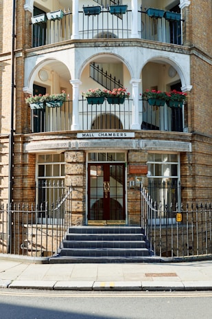 A multi-story building with arched balconies containing flower pots with red flowers, black railings, and a grand entrance with a staircase leading up to double glass doors. The building is made of light-colored brick and features a sign reading 'Mall Chambers.' The railings and the main door are framed with decorative metalwork.