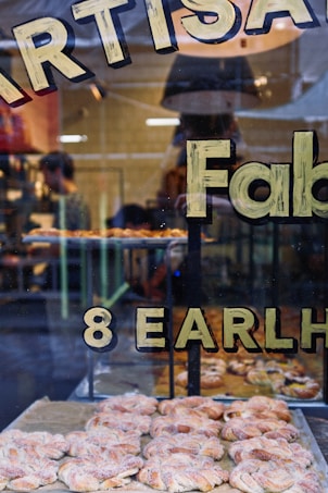 A bakery display window featuring freshly baked pastries arranged on trays. The window has bold lettering that partially reads 'ARTIS...' and '8 EARLH...'. Inside, a person is working with shelves or equipment in the background, adding to the busy atmosphere.