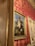 An ornate interior setting featuring a classical painting of a floral arrangement in a vase. The painting, framed in gold, is hung on a wall covered with red and gold damask wallpaper. The scene exudes elegance with the intricate design elements and rich color palette.