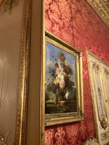 An ornate interior setting featuring a classical painting of a floral arrangement in a vase. The painting, framed in gold, is hung on a wall covered with red and gold damask wallpaper. The scene exudes elegance with the intricate design elements and rich color palette.