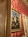 An ornate interior setting featuring a classical painting of a floral arrangement in a vase. The painting, framed in gold, is hung on a wall covered with red and gold damask wallpaper. The scene exudes elegance with the intricate design elements and rich color palette.