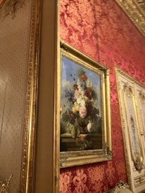 An ornate interior setting featuring a classical painting of a floral arrangement in a vase. The painting, framed in gold, is hung on a wall covered with red and gold damask wallpaper. The scene exudes elegance with the intricate design elements and rich color palette.