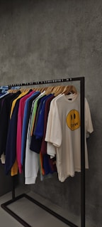 A clothing rack displaying various t-shirts and sweatshirts on hangers against a textured grey wall. The shirts are in assorted colors including dark blue, pink, yellow, green, white, and beige. The beige shirt in the front features a smiley face graphic with the word 'drew' below it.