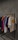 A clothing rack displaying various t-shirts and sweatshirts on hangers against a textured grey wall. The shirts are in assorted colors including dark blue, pink, yellow, green, white, and beige. The beige shirt in the front features a smiley face graphic with the word 'drew' below it.