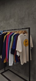 A clothing rack displaying various t-shirts and sweatshirts on hangers against a textured grey wall. The shirts are in assorted colors including dark blue, pink, yellow, green, white, and beige. The beige shirt in the front features a smiley face graphic with the word 'drew' below it.