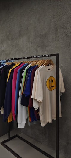 A clothing rack displaying various t-shirts and sweatshirts on hangers against a textured grey wall. The shirts are in assorted colors including dark blue, pink, yellow, green, white, and beige. The beige shirt in the front features a smiley face graphic with the word 'drew' below it.