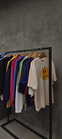 A clothing rack displaying various t-shirts and sweatshirts on hangers against a textured grey wall. The shirts are in assorted colors including dark blue, pink, yellow, green, white, and beige. The beige shirt in the front features a smiley face graphic with the word 'drew' below it.
