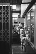 A grocery market setting with stacks of various goods and two individuals interacting. The surroundings include shelves filled with items and a mix of architectural elements like metal railings and glass. Soft lighting illuminates the area, creating a contrast between light and shadow.